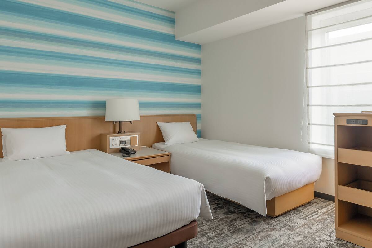 Habitación de hotel con dos camas, una mesita de noche con lámpara, una pequeña mesa y una ventana con cortinas translúcidas.