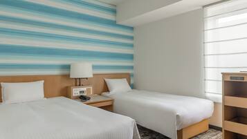 A hotel room with two beds, a nightstand with a lamp, a small table, and a window with sheer curtains.