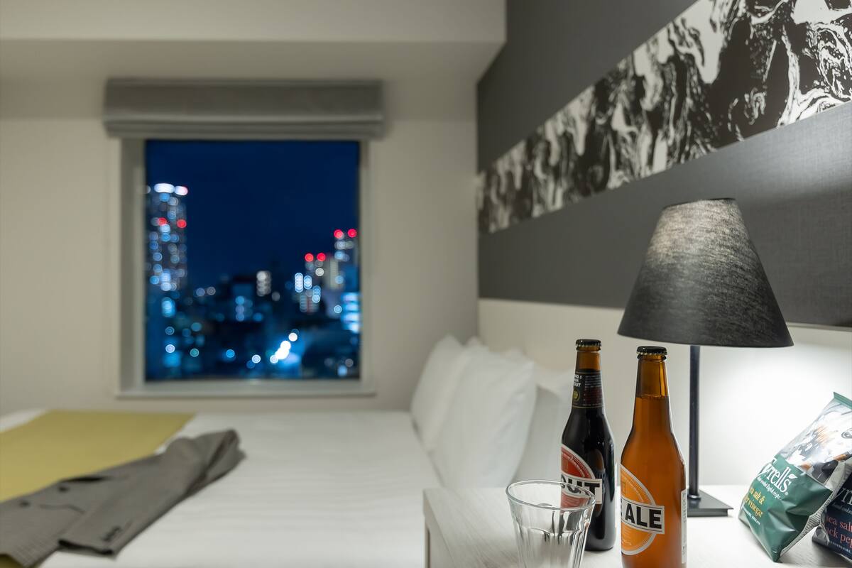 Habitación de hotel con cama, ventana con vista a la ciudad, lámpara y dos botellas de cerveza.