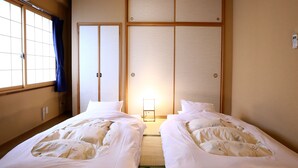 Een hotelkamer met twee eenpersoonsbedden, een schuifdeur van shoji-papier, een bedlampje en uitzicht naar buiten door een raam met een blauw gordijn.
