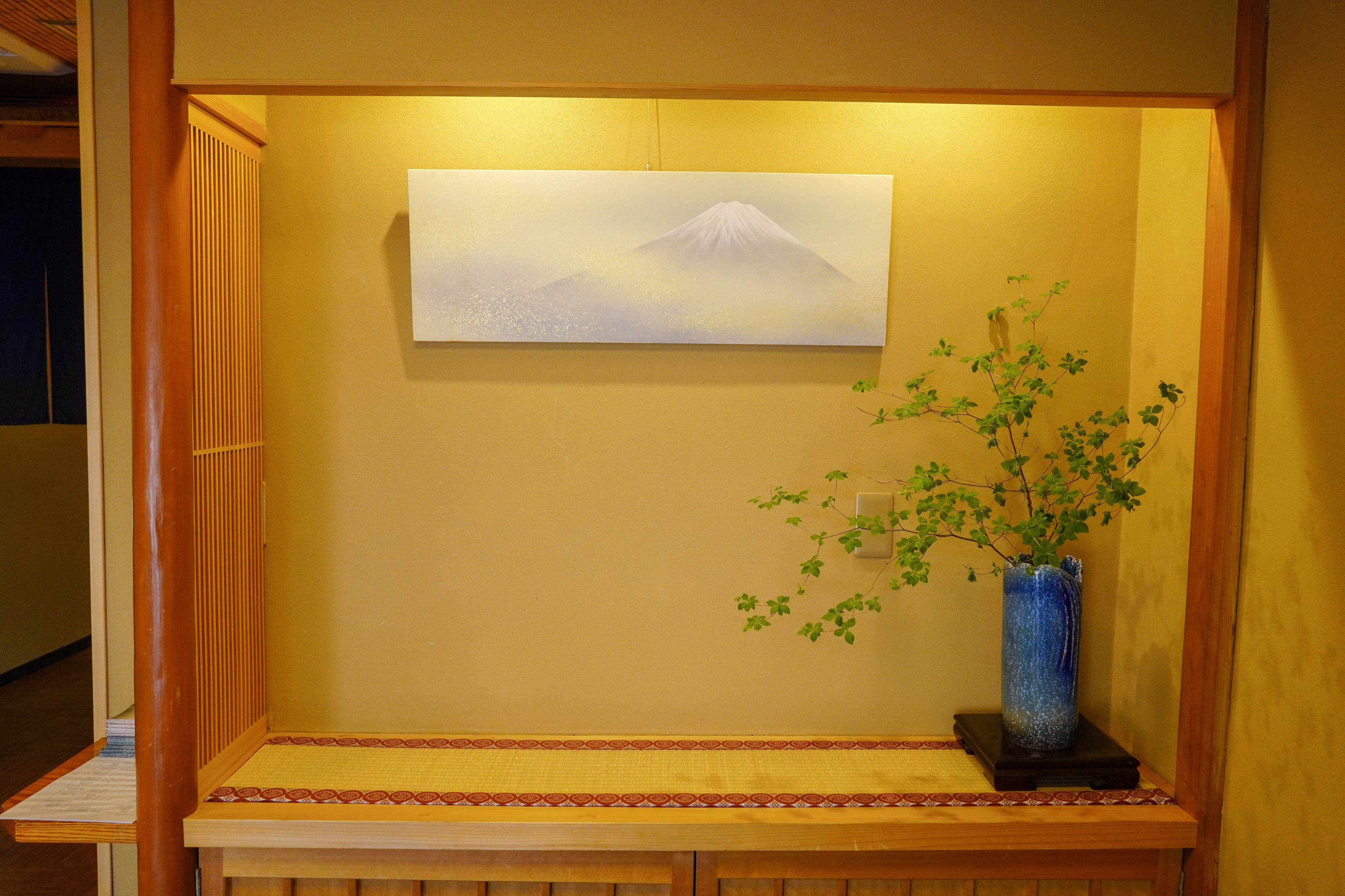 A room with a wall-mounted painting of a mountain, a small table with a vase of green plants, and a wooden bench.