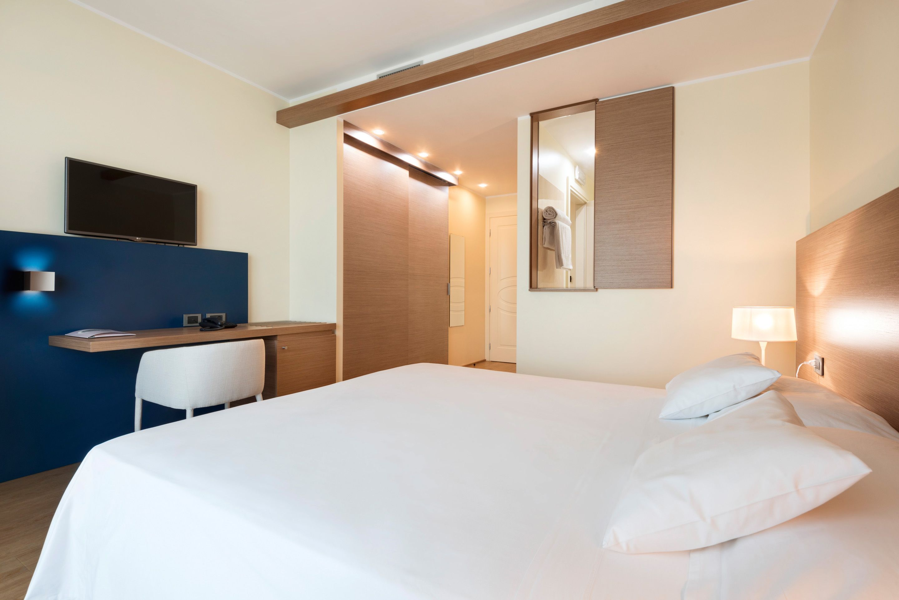 A hotel room with a bed, a desk, a chair, a TV, a mirror, and a door.
