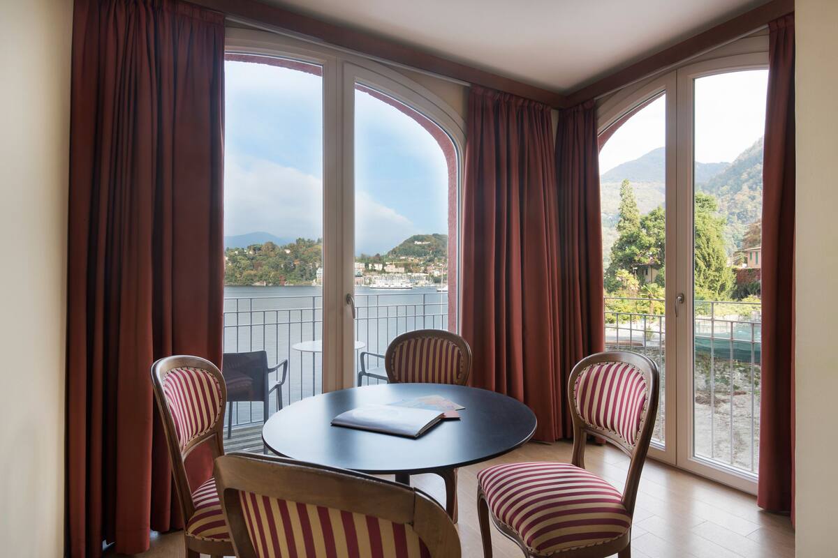 A room with a round table and four striped chairs, large arched windows with curtains, and a view of a hillside.