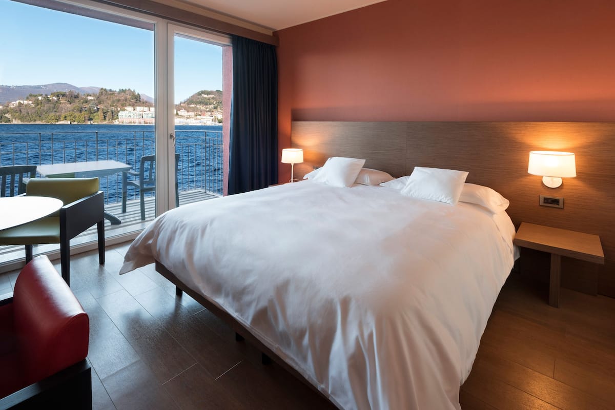 A hotel room with a large bed, two bedside tables with lamps, a small table and chair, and a view of the sea and hills.