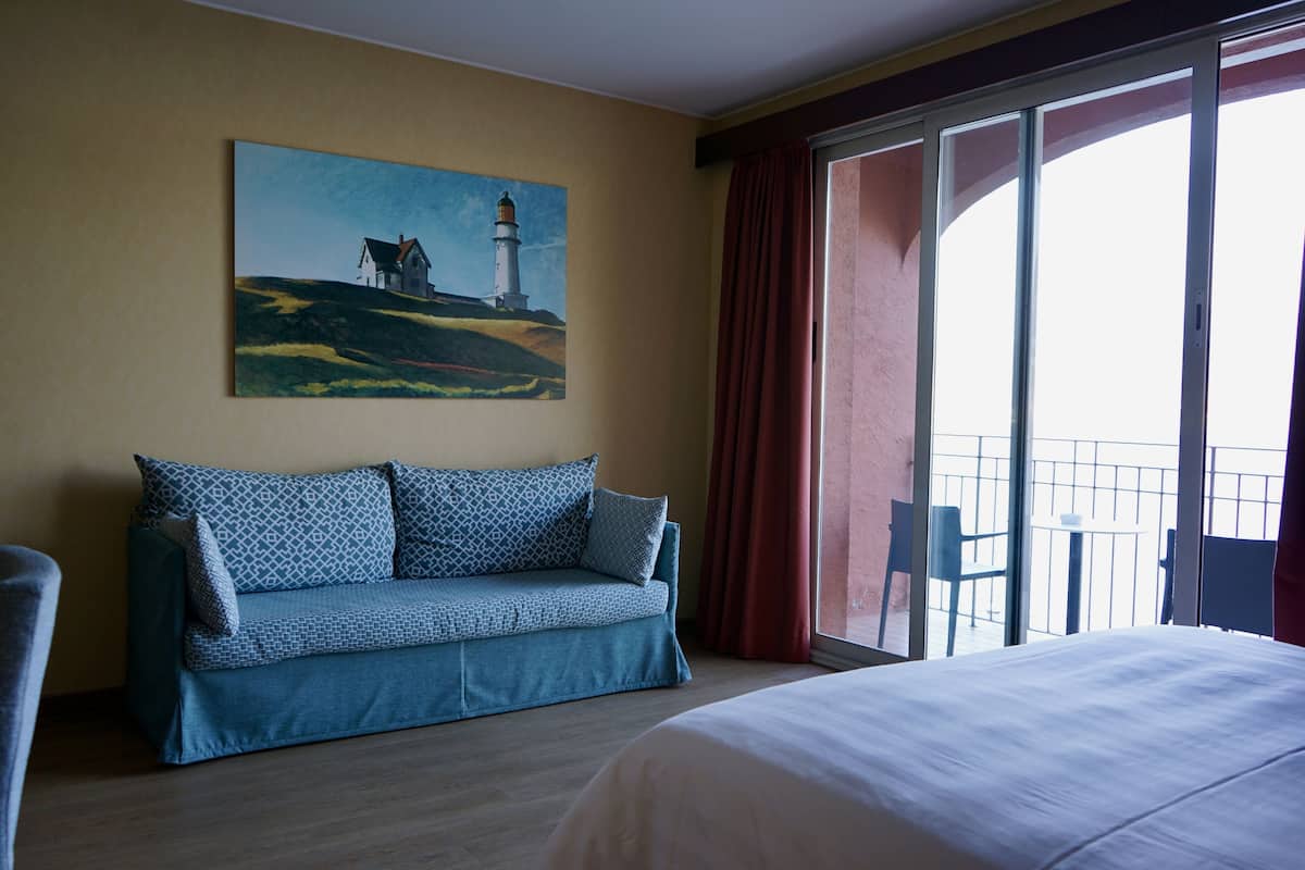 A hotel room with a large bed, a blue sofa with patterned cushions, a painting of a lighthouse, and a balcony with chairs.