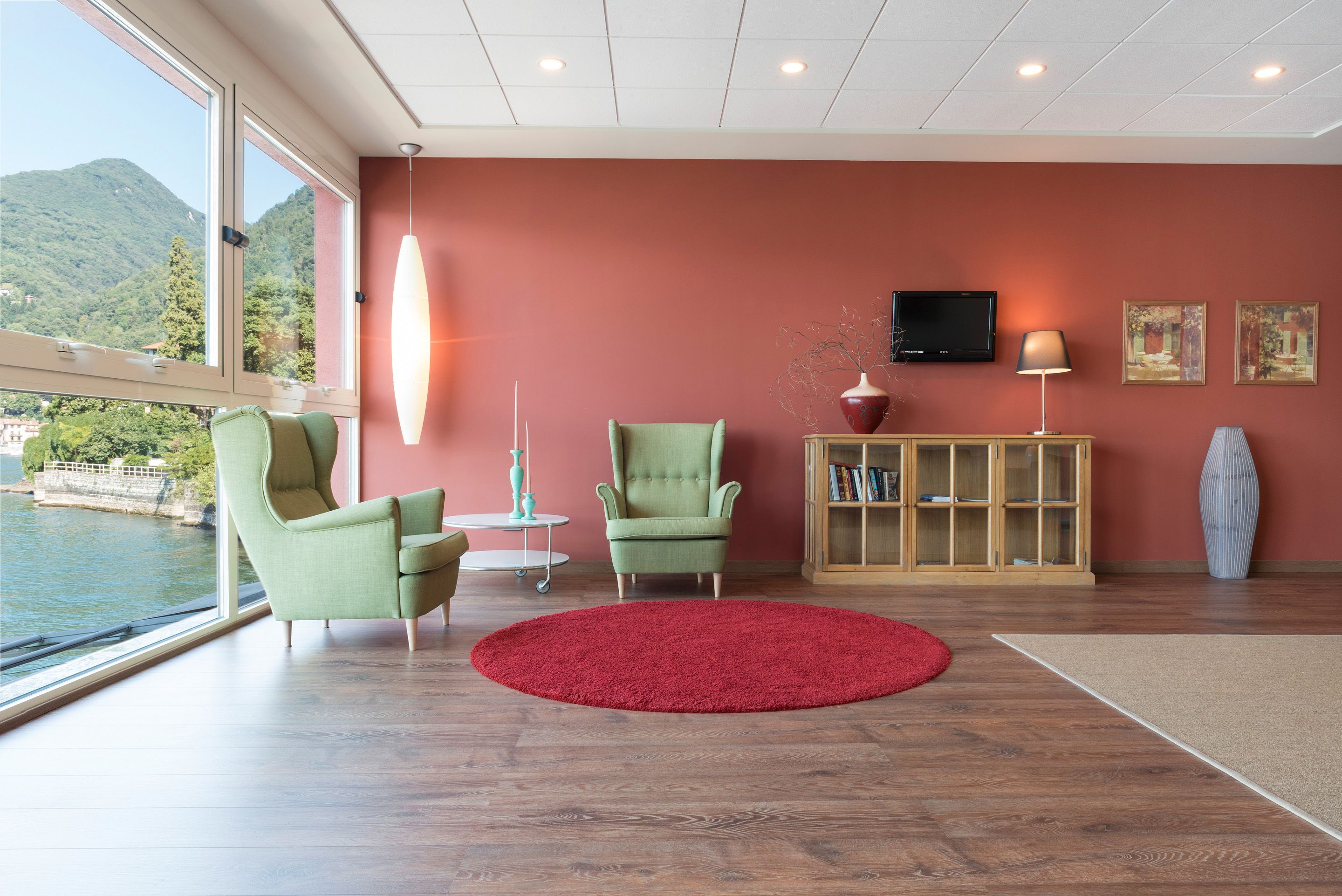 A modern living room with a large window overlooking a body of water and hills, featuring two green armchairs, a red rug, a wooden cabinet, and a television.