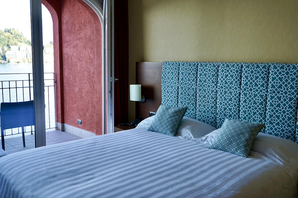 A hotel room with a bed, a nightstand, a lamp, a chair, and a view of the outdoors.