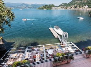 A terrace with outdoor seating overlooking a lake, a dock with a boat, and a clear view of the water and distant hills. - Il Porticciolo (Laveno Mombello)