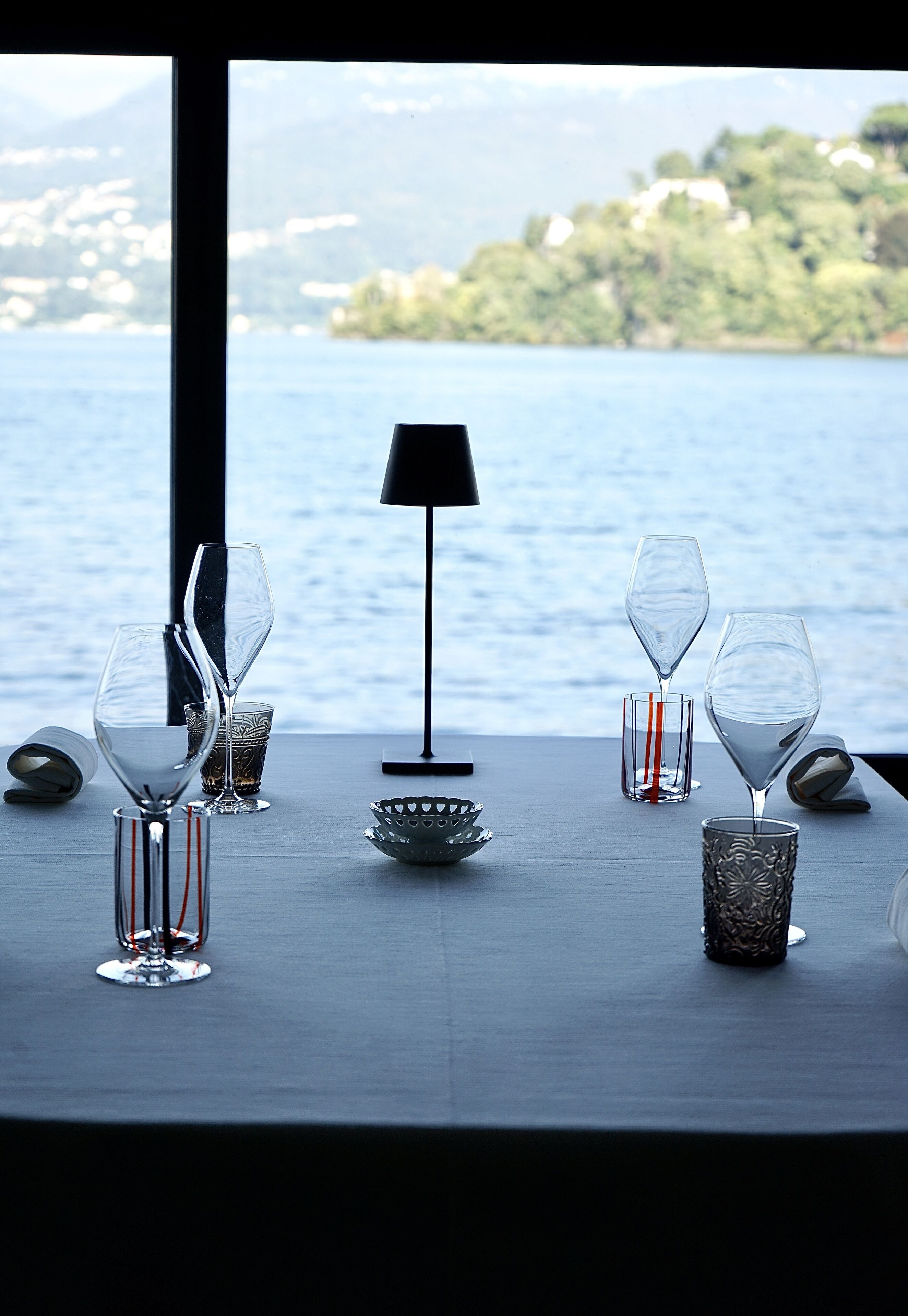 A table set with wine glasses and a lamp, overlooking a body of water with a view of a distant shoreline.