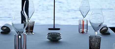 A table set with wine glasses and a lamp, overlooking a body of water with a view of a distant shoreline.