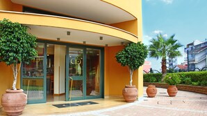 L’entrée d’un hôtel, avec une façade jaune incurvée, de grands bacs en terre cuite ornés d’arbres et un ciel dégagé.