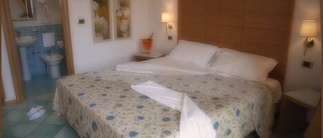 A bedroom with a bed, a nightstand, a wall-mounted lamp, a painting, a bathroom with a sink and mirror, and a pair of slippers on the floor.
