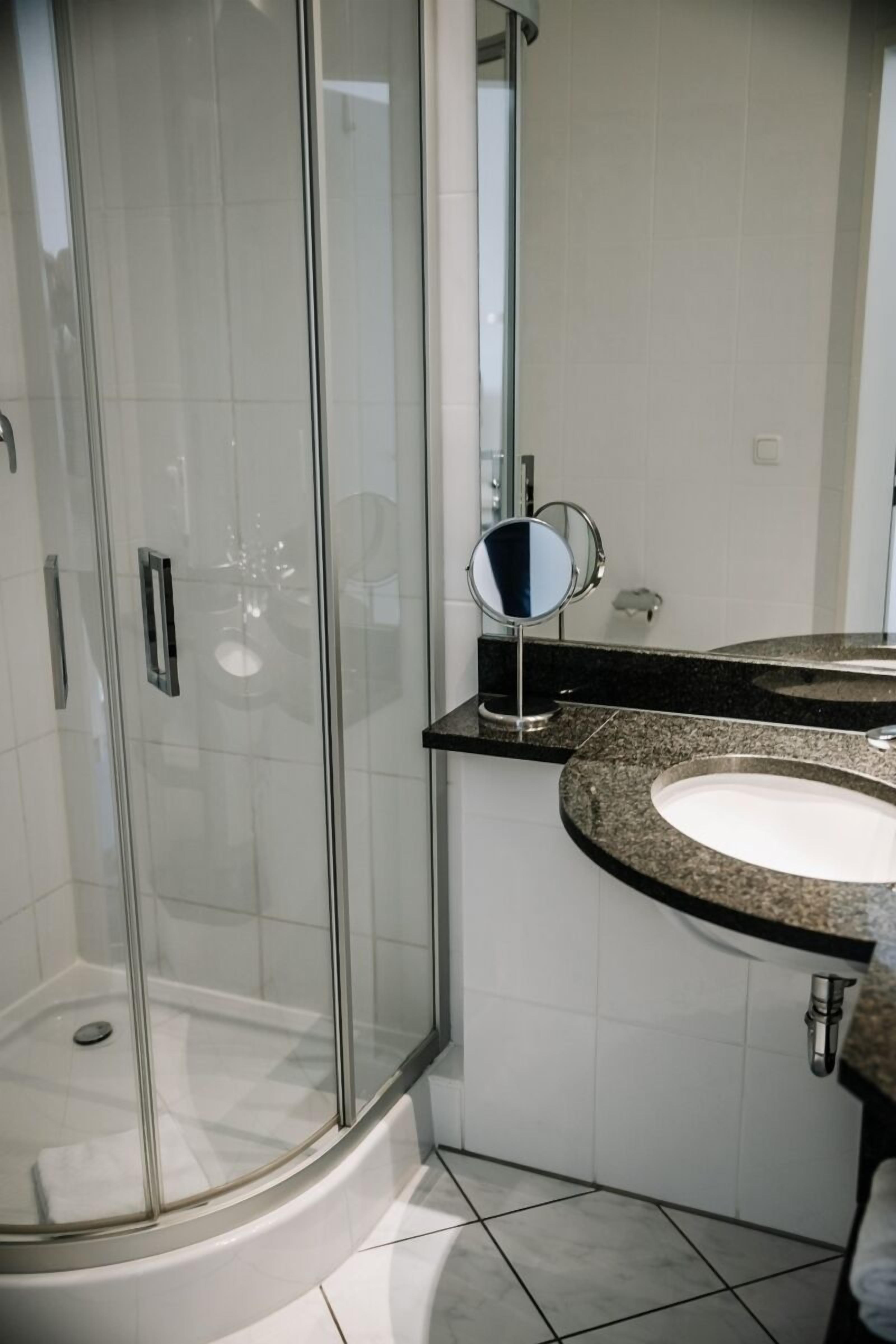 A modern bathroom with a glass-enclosed shower, a round mirror, and a granite countertop sink.
