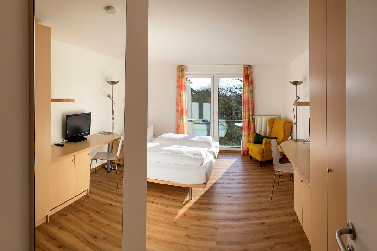 A modern hotel room with a wooden floor, a bed with white bedding, a desk with a TV, a chair, a sofa, and a window with curtains.