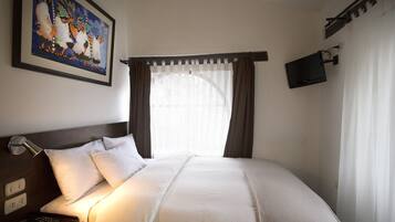 A hotel room with a bed, a nightstand with a lamp, a painting on the wall, a TV mounted on the wall, and a window with curtains.