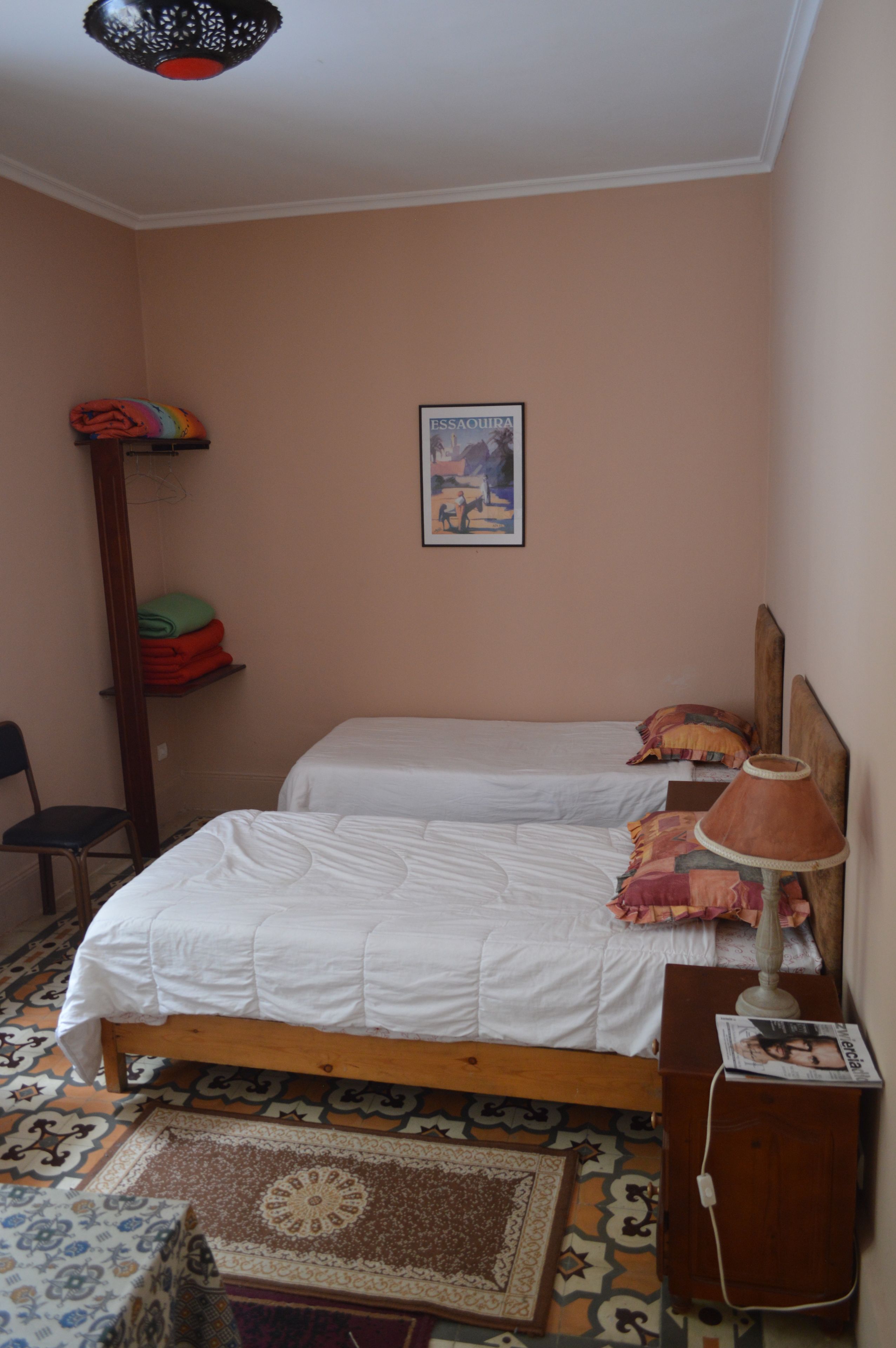 Une chambre avec deux lits, une chaise, une table, une lampe et un tableau encadré au mur.