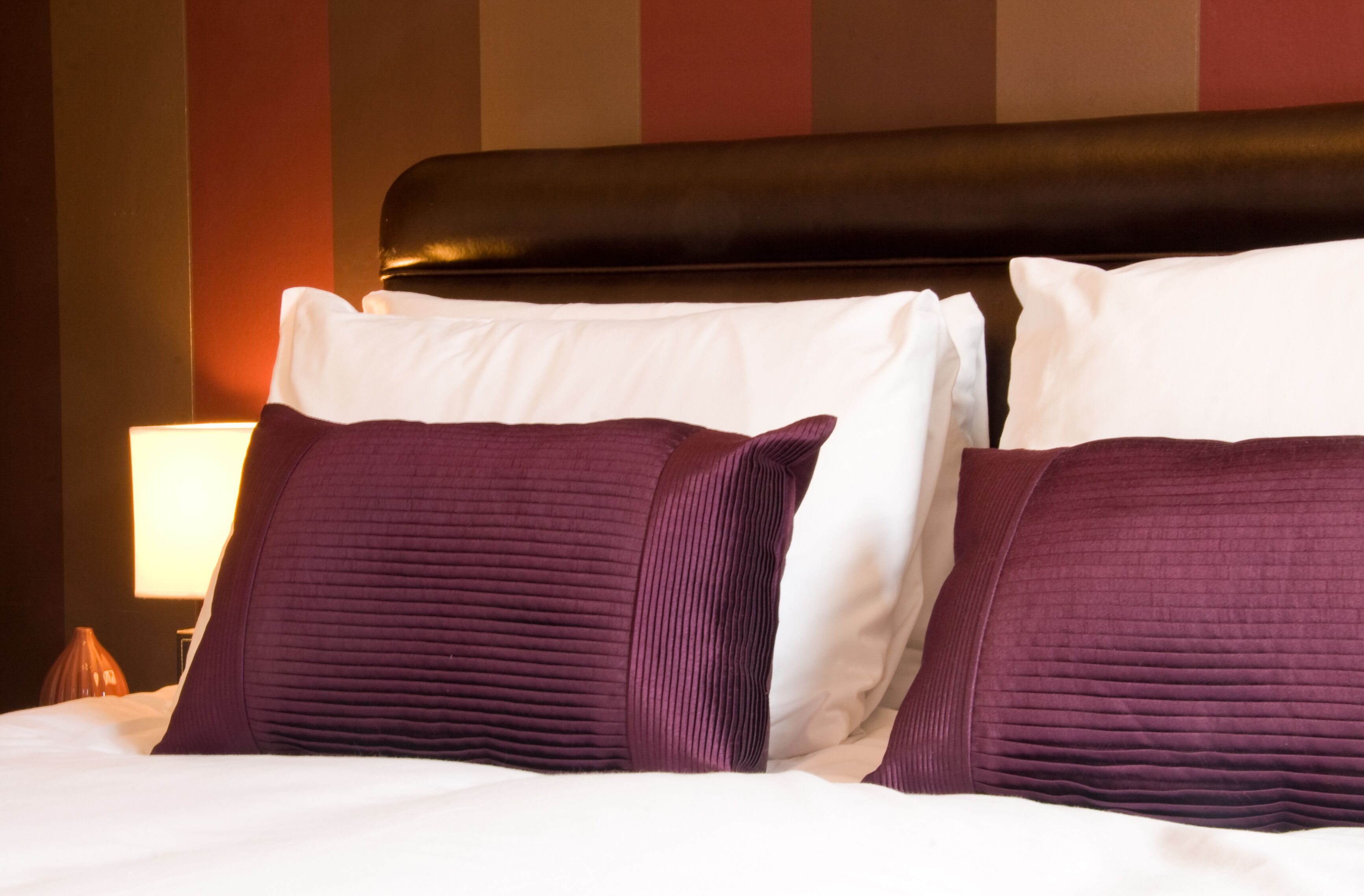 A neatly made bed with white and purple pillows, a black headboard, and a lamp on a bedside table.
