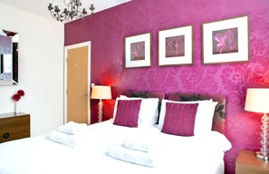 A hotel room with a large bed, a wooden nightstand, a mirror, and a wall with three framed pictures. - Harrogate Boutique Apartments (Harrogate)