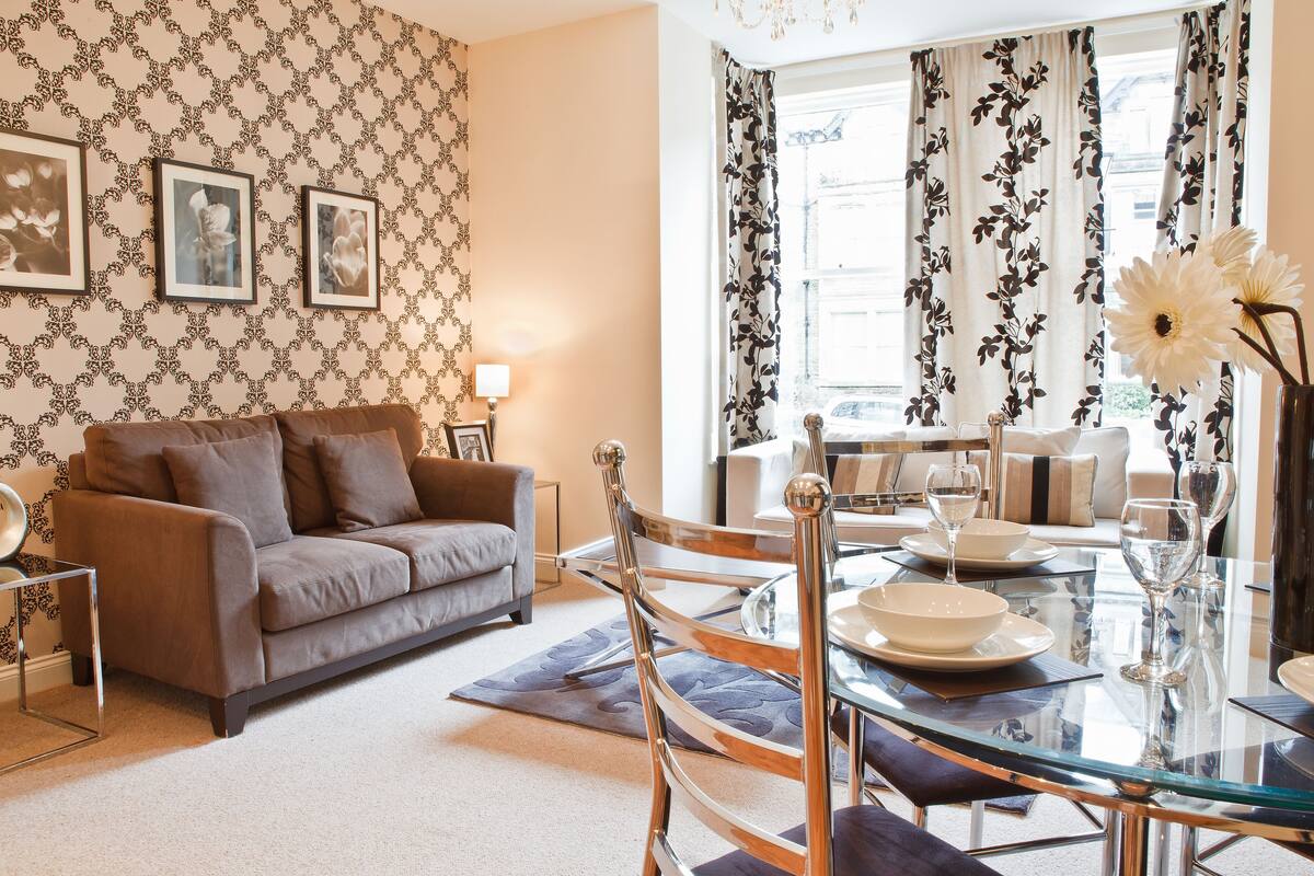A living room with a brown sofa, a glass dining table set for four, and a patterned wallpaper.