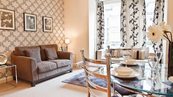 A living room with a brown sofa, a glass dining table set for four, and a patterned wallpaper.