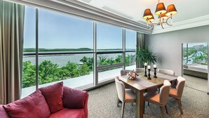 Une chambre d’hôtel moderne dotée d’une grande fenêtre donnant sur une rivière et un paysage verdoyant. On y trouve un coin repas avec une table et des chaises, ainsi qu’un canapé rouge.
