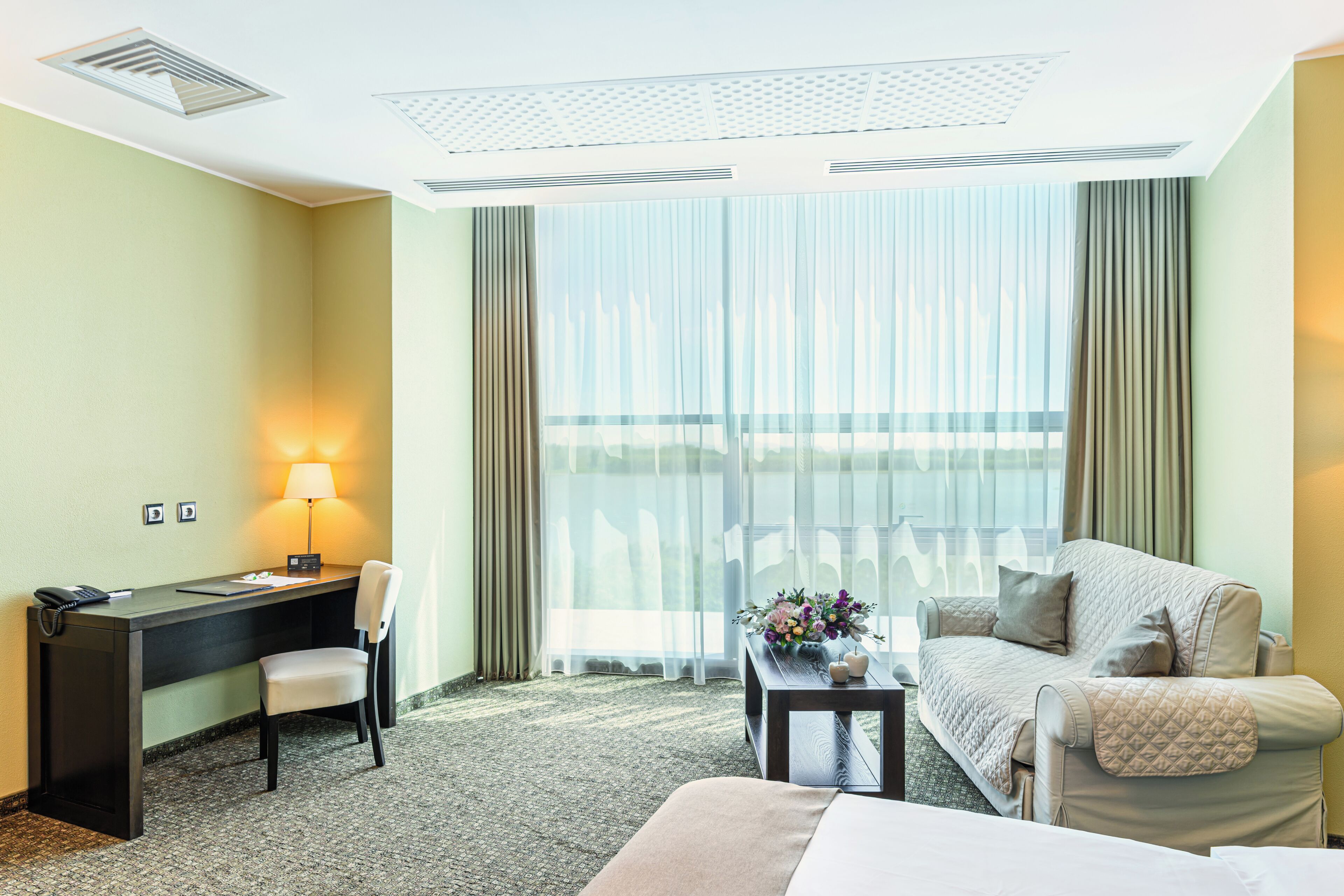 Ein Hotelzimmer mit Schreibtisch, Stuhl, Bett und einem großen Fenster mit Vorhängen.