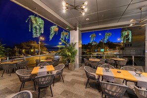 An outdoor dining area with wicker chairs and tables, decorated with hanging plant lights, and overlooking a street at night. - Vega Hotel (Galati)