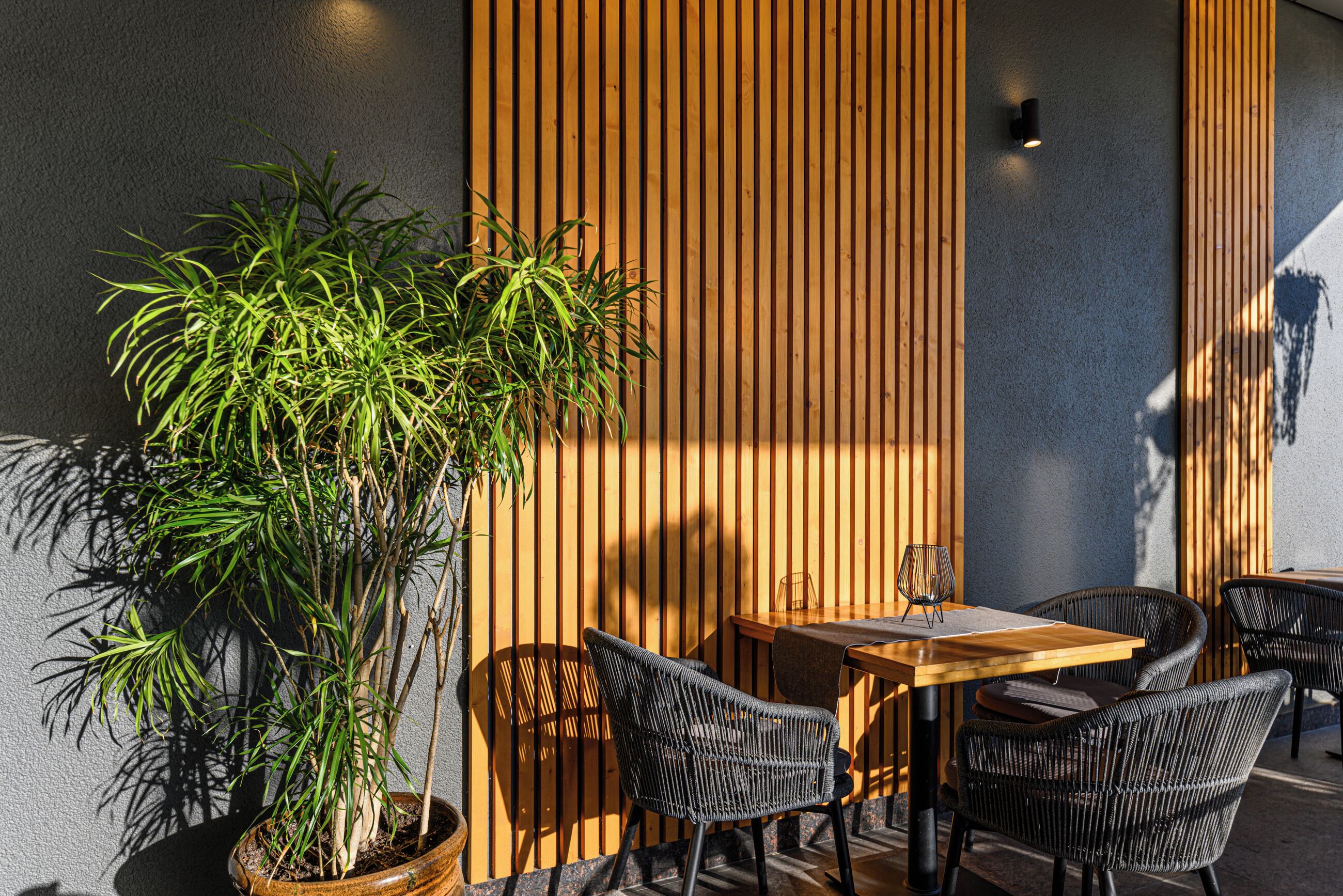 An outdoor seating area with a wooden table and chairs, a potted plant, and a vertical wooden slat wall.