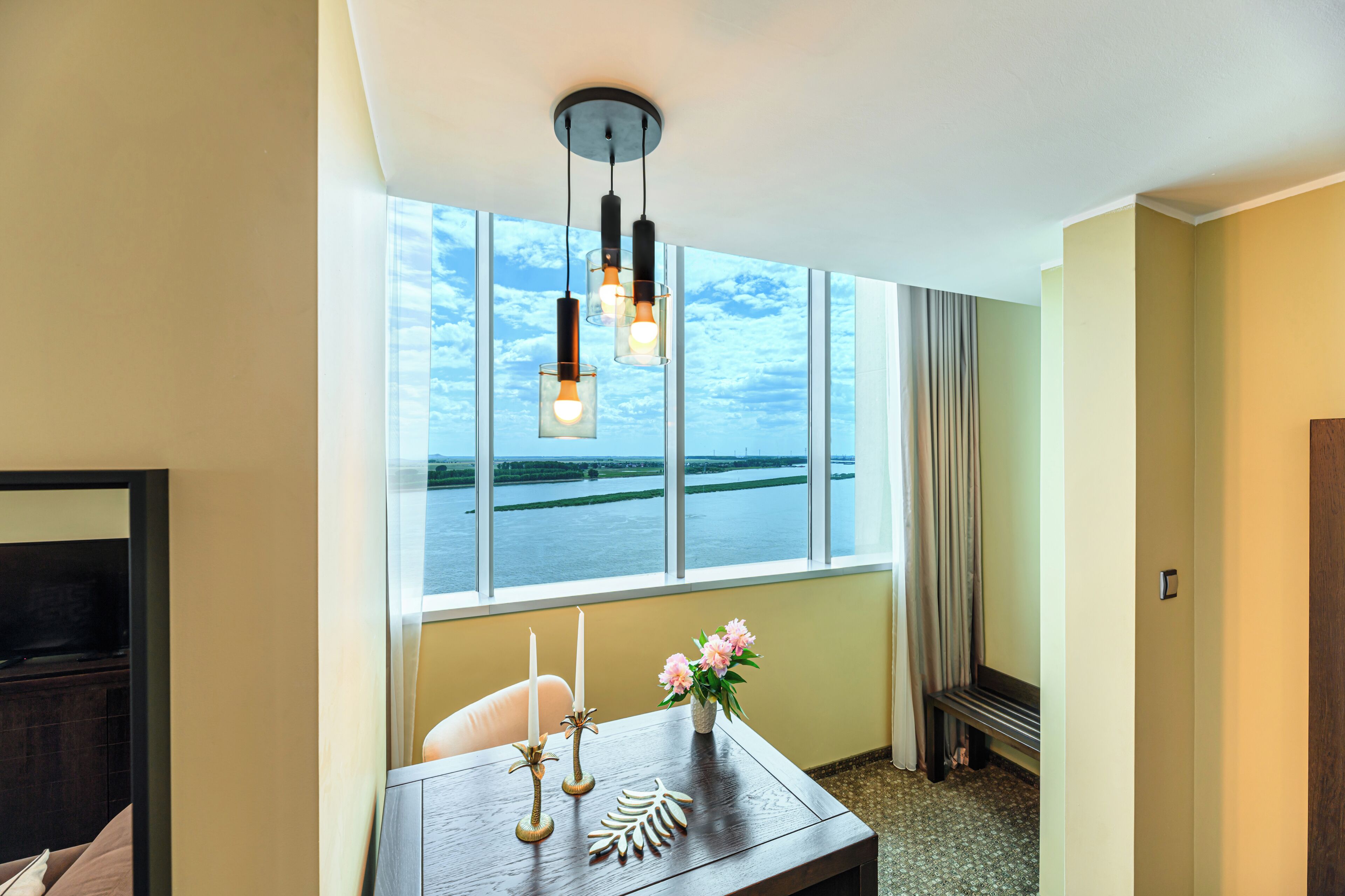 Una habitación de hotel con un ventanal grande, una mesa de madera con velas y flores, y un banco.