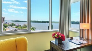 Une chambre d’hôtel dotée d’une grande fenêtre donnant sur une rivière et sur des bâtiments éloignés. On y trouve un bureau avec une lampe, un vase de fleurs et un bloc-notes.