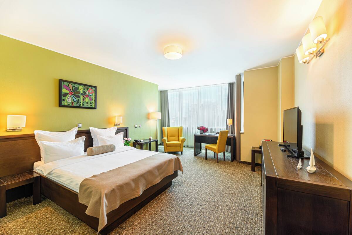 A hotel room with a large bed, a desk, a chair, a TV, and a painting on the wall.
