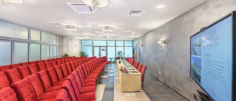 A conference room with red chairs arranged in rows, a long table with a laptop, and a large screen displaying text.