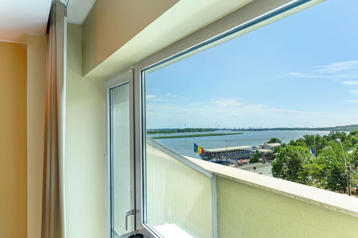 A room with a large window overlooking a waterfront view, including a flag, buildings, and greenery.