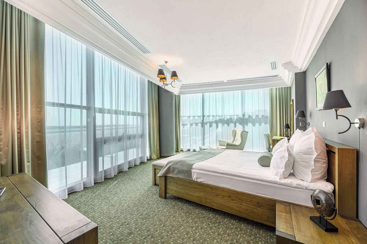 A hotel room with a large bed, a wooden nightstand, a desk, and a view of the city through large windows.