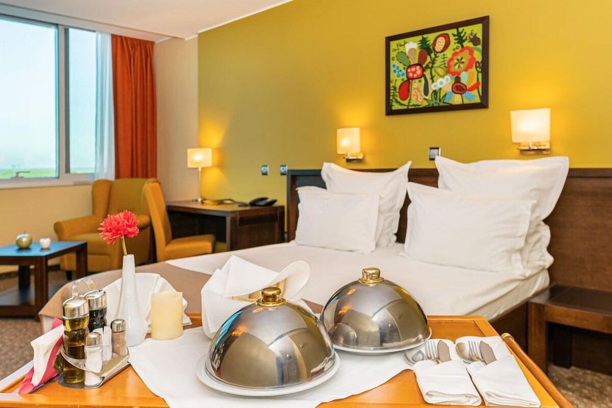 A hotel room with a bed, bedside lamps, a desk, a chair, a window with curtains, and a painting on the wall.