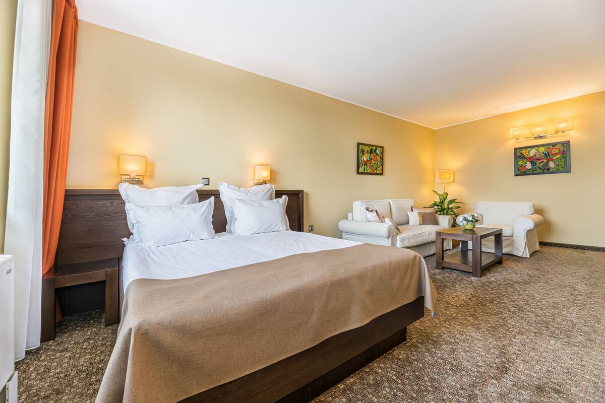 A hotel room with a large bed, two white armchairs, a small table, and two wall-mounted lamps.