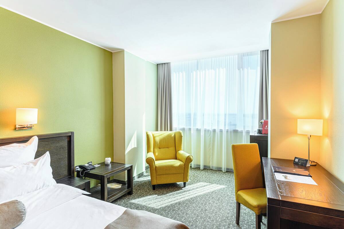 A hotel room with a bed, a desk, a chair, a lamp, and a window with curtains.