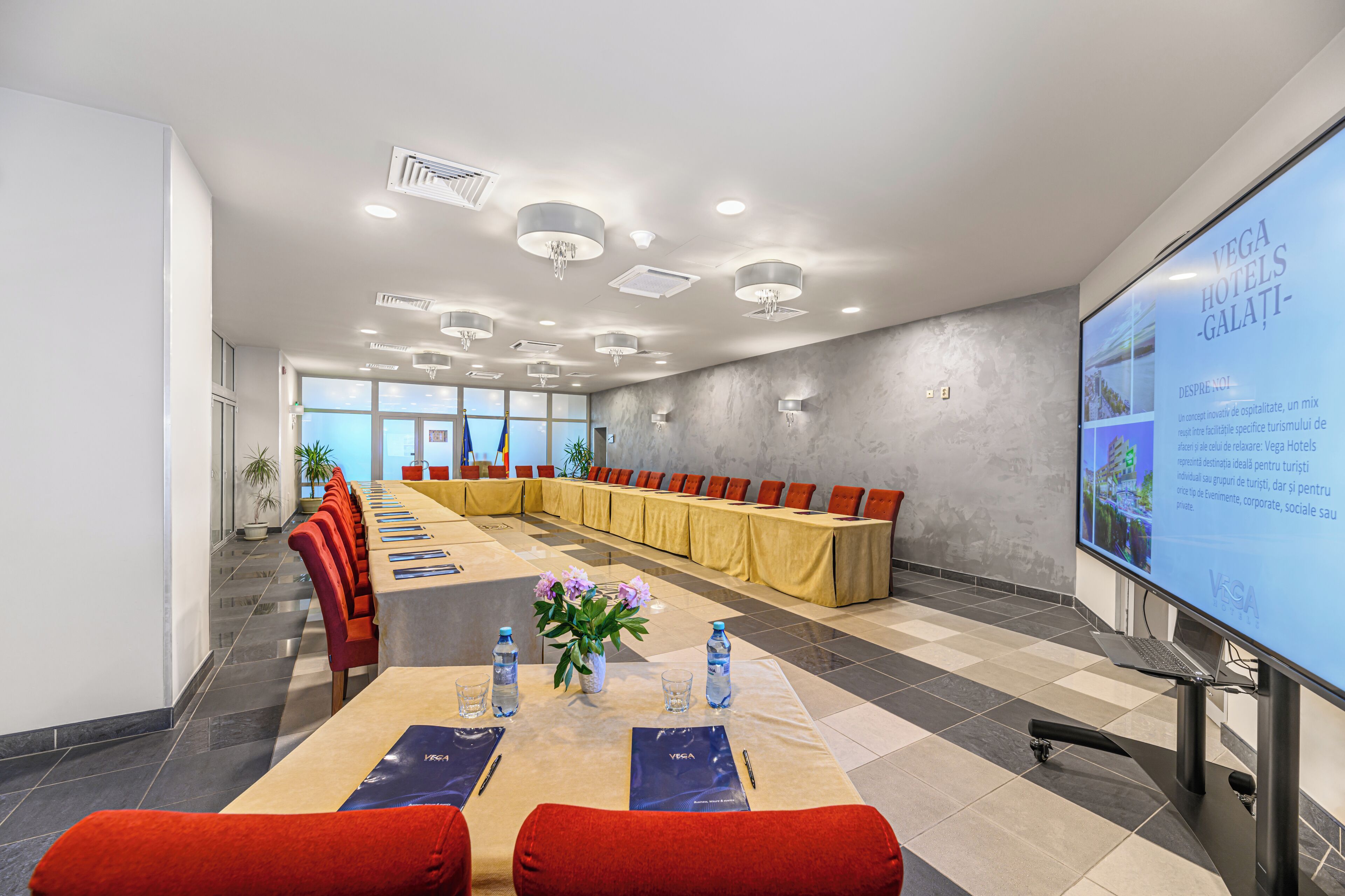 Una sala de conferencias con una mesa larga, sillas rojas y una pantalla de proyector mostrando información del hotel.