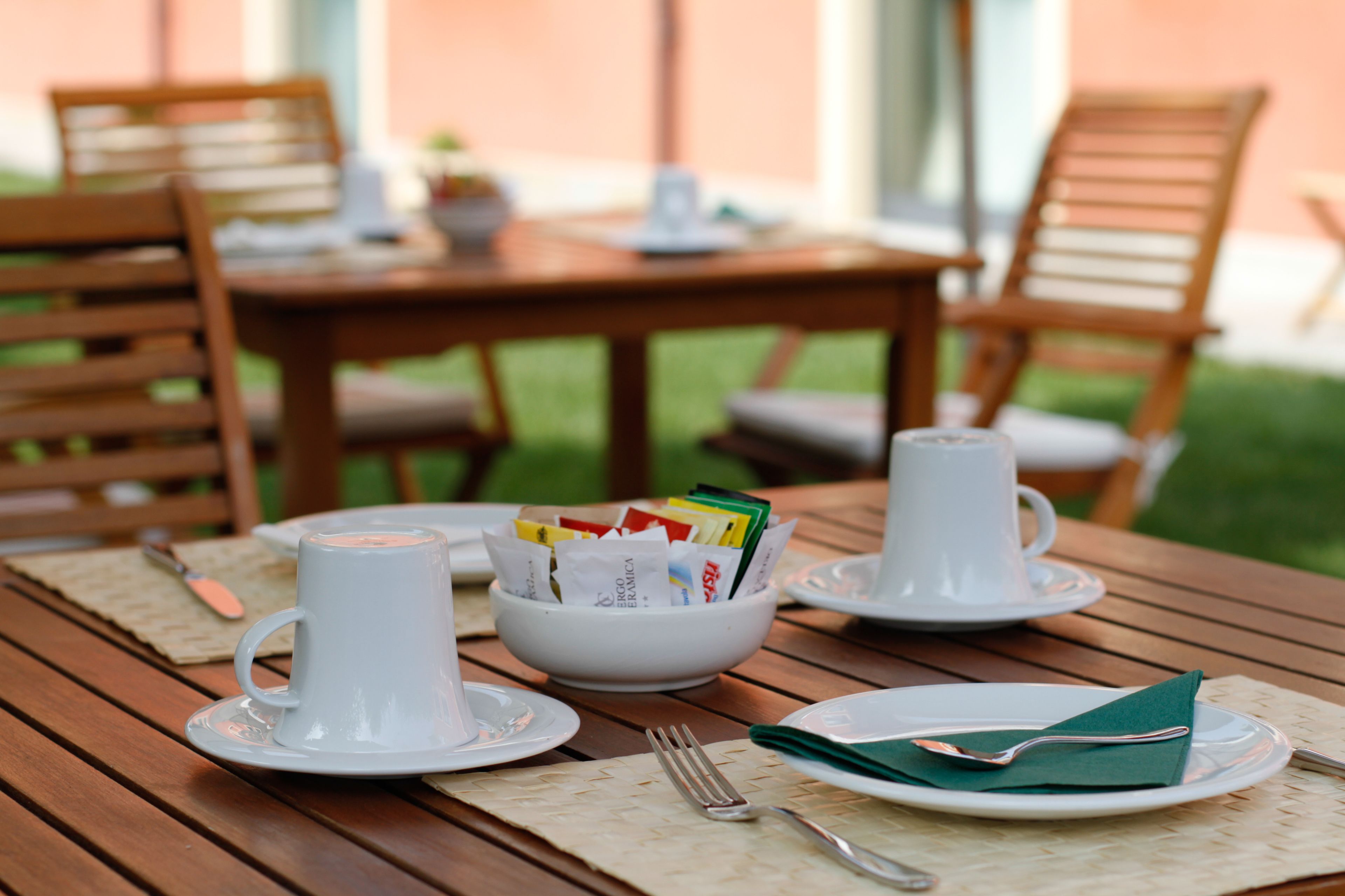 A wooden dining table set with white cups, saucers, a bowl of magazines, and a folded green napkin.
