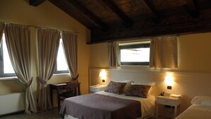 A hotel room with a large bed, two bedside tables, a desk, and a chair. There are curtains on the windows, and the room has a wooden ceiling.