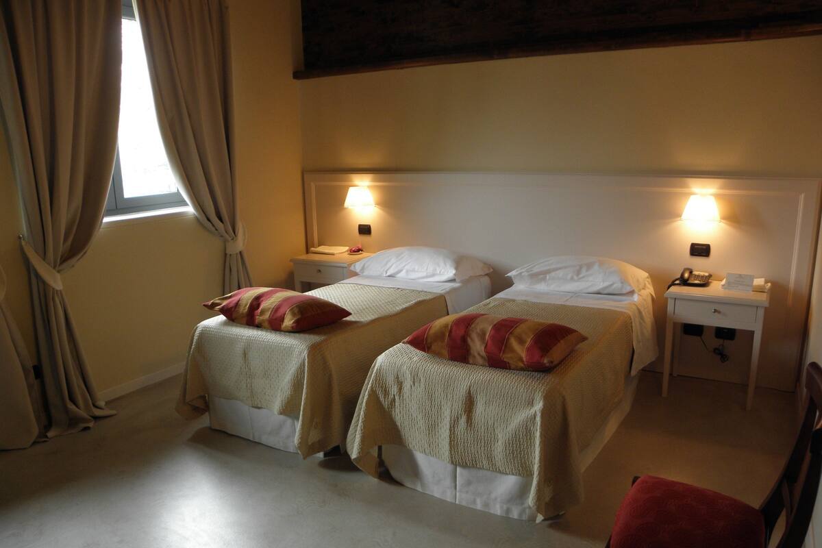 A hotel room with two beds, each with a pillow and a bedside table with a lamp.