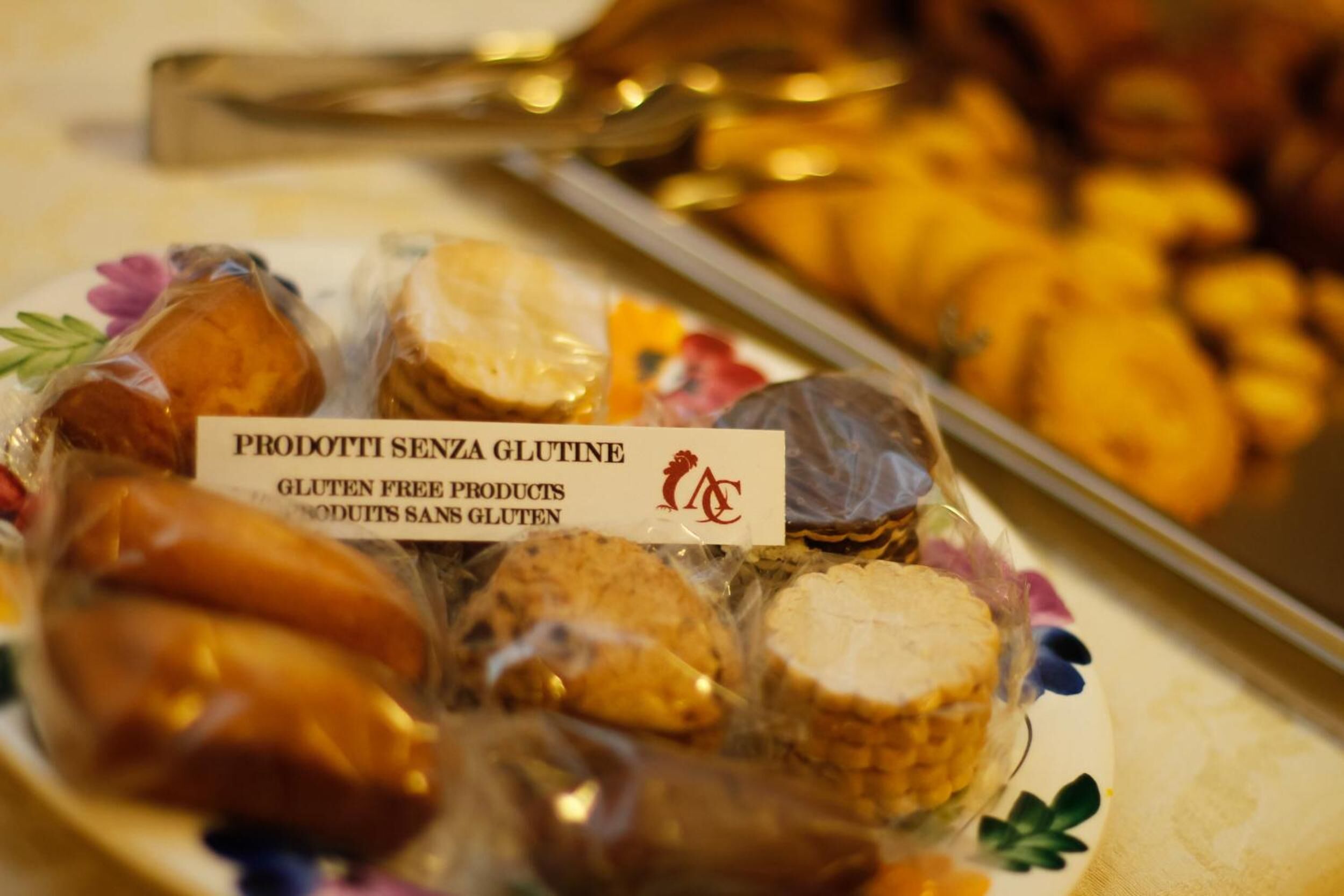 A plate of gluten-free products labeled "PRODOTTI SENZA GLUTINE" with a tray of pastries in the background.