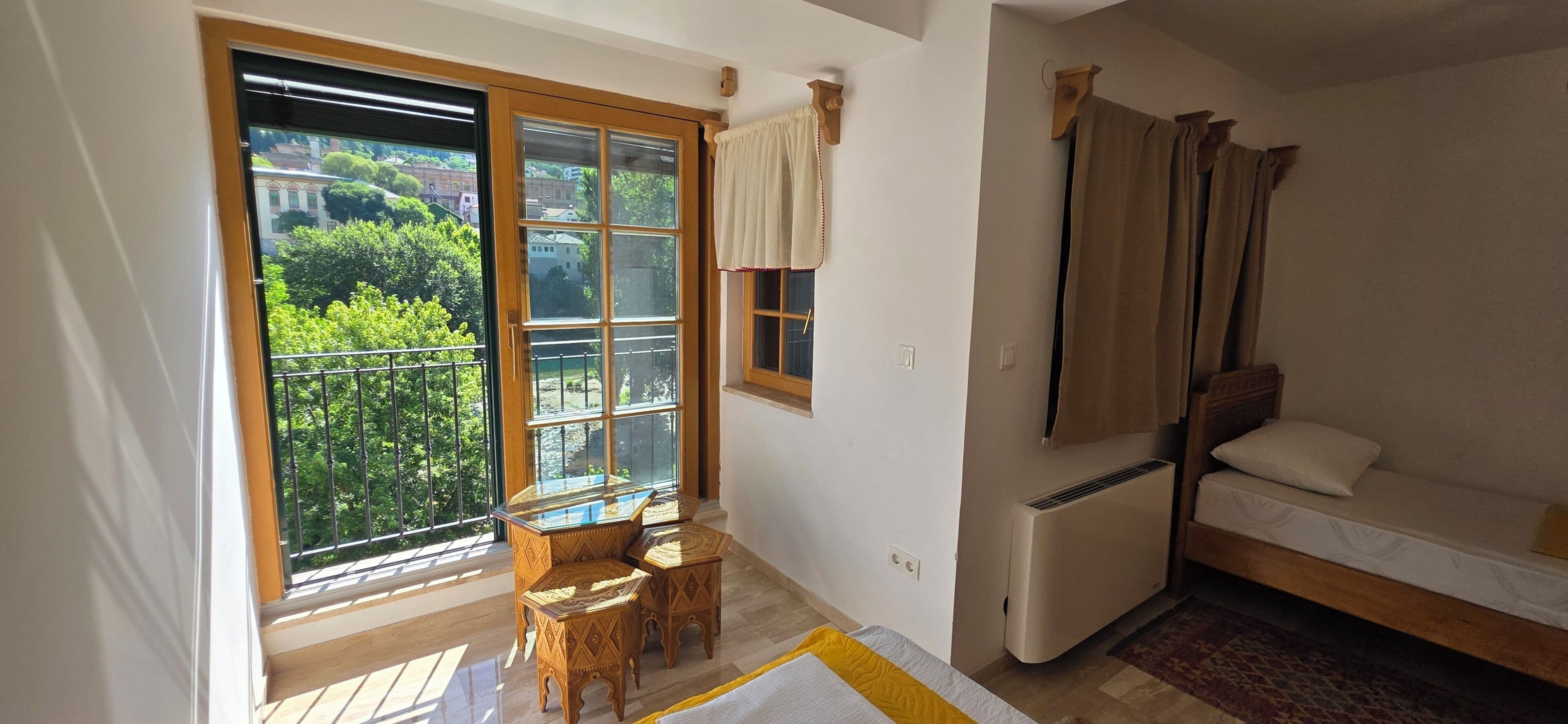 A room with a balcony, a bed, a table with chairs, and a window with curtains.
