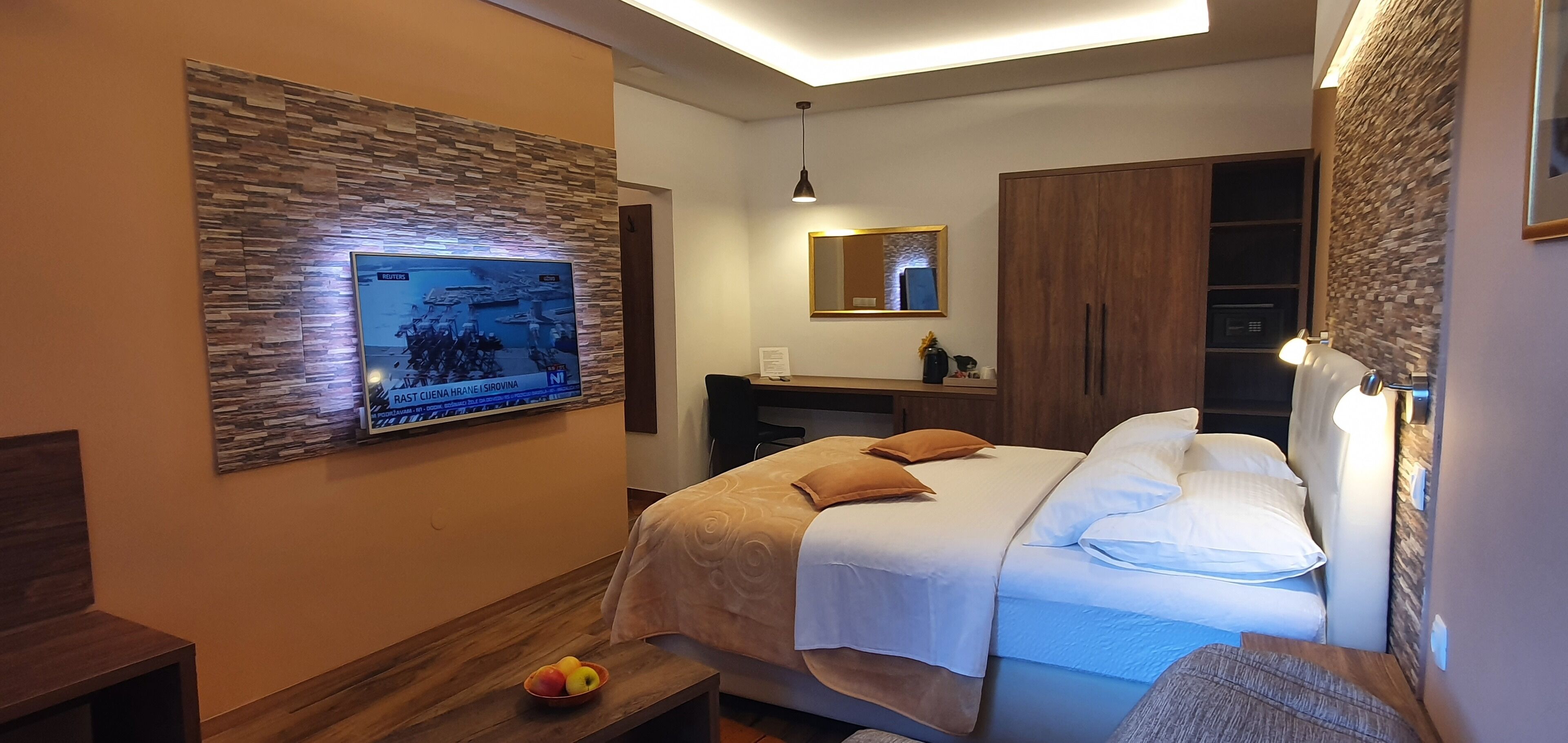 A modern hotel room with a large flat-screen TV mounted on the wall, a bed with white bedding and two pillows, a wooden nightstand with a bowl of fruit, a mirror above the bed, and a wardrobe with a mirror.