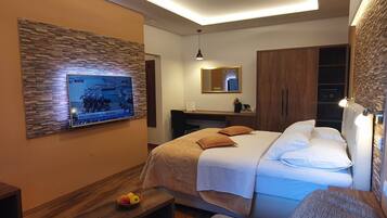 A modern hotel room with a large flat-screen TV mounted on the wall, a bed with white bedding and two pillows, a wooden nightstand with a bowl of fruit, a mirror above the bed, and a wardrobe with a mirror.