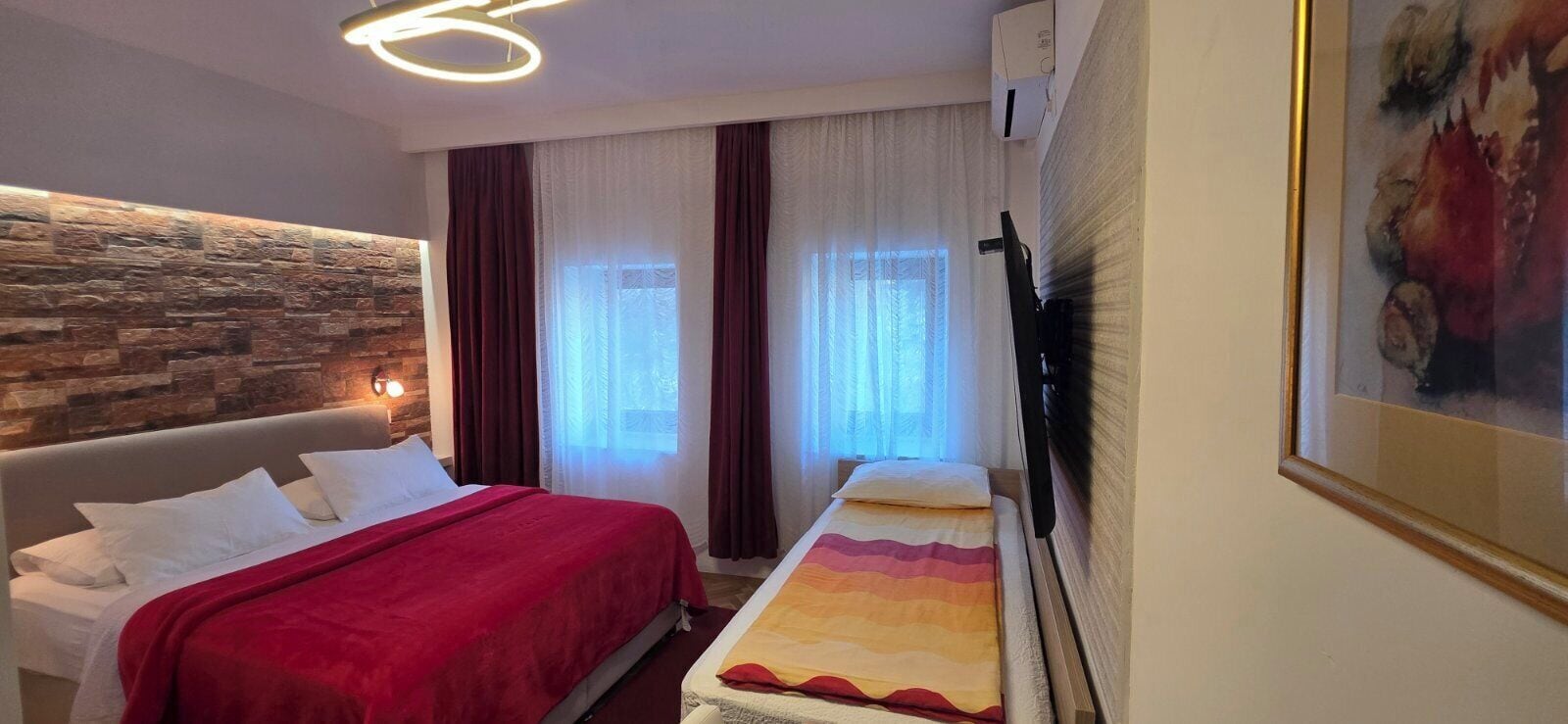 A hotel room with a stone accent wall, a bed with a red blanket, a bedside table, a TV, and a painting on the wall.