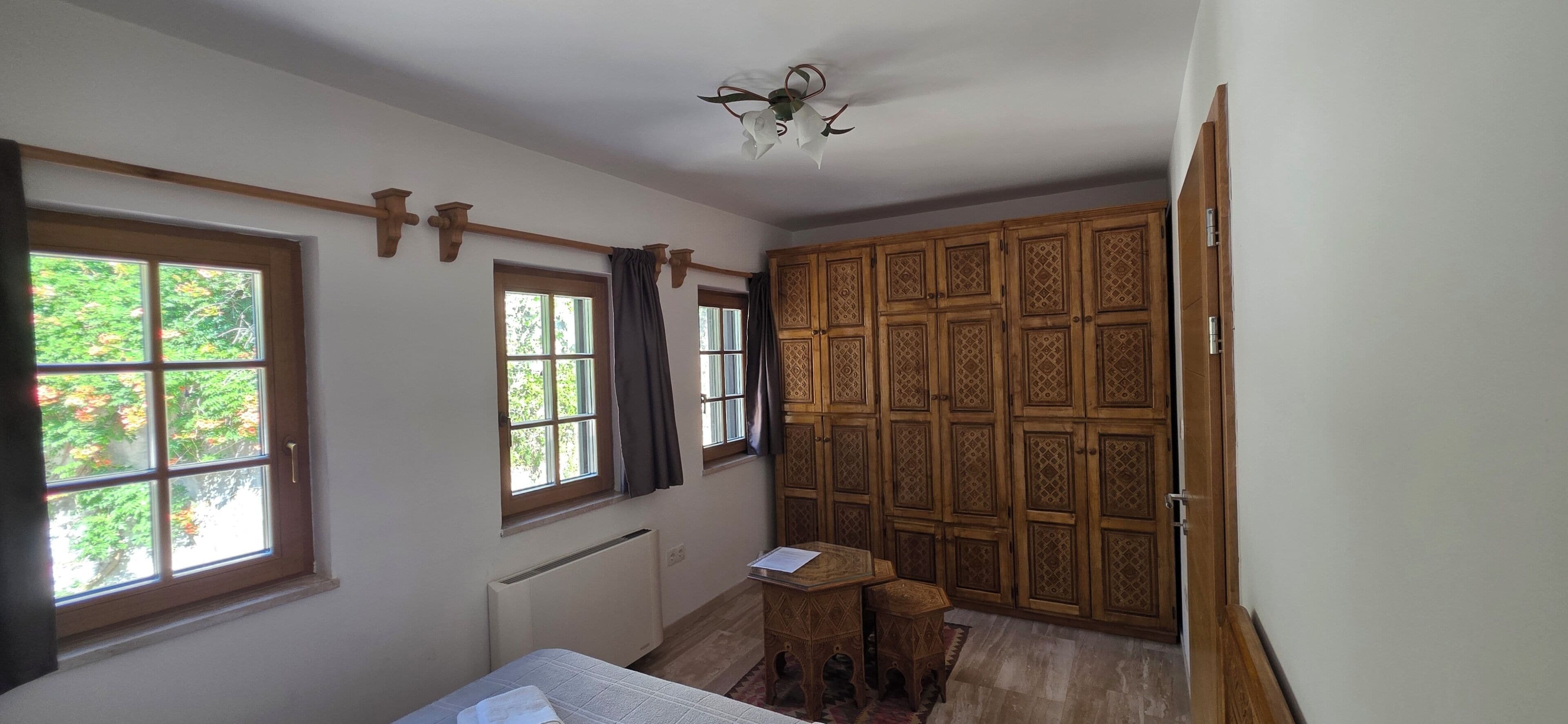 Una habitació amb un llit, una taula petita, un radiador, una finestra amb cortines i un armari de fusta.