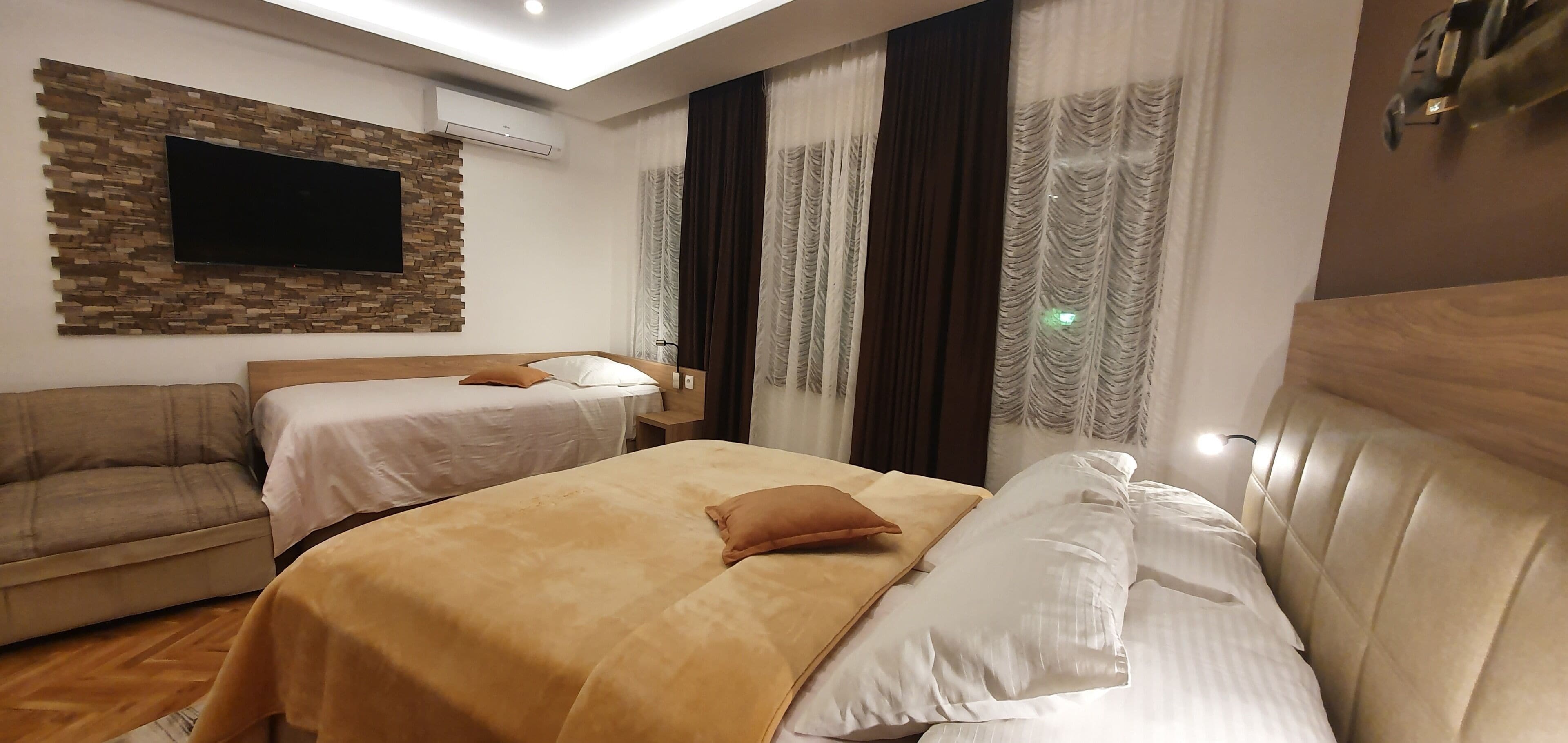 A hotel room with two beds, a flat-screen TV mounted on a stone wall, a wooden headboard, and a window with curtains.