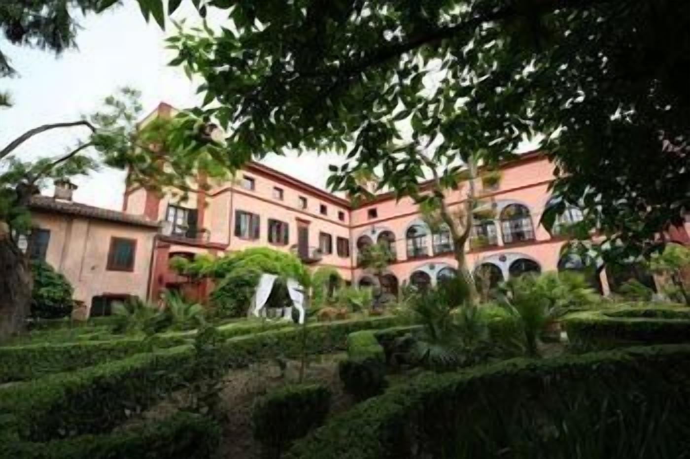 A building with multiple floors, arched windows, and a courtyard with manicured hedges.
