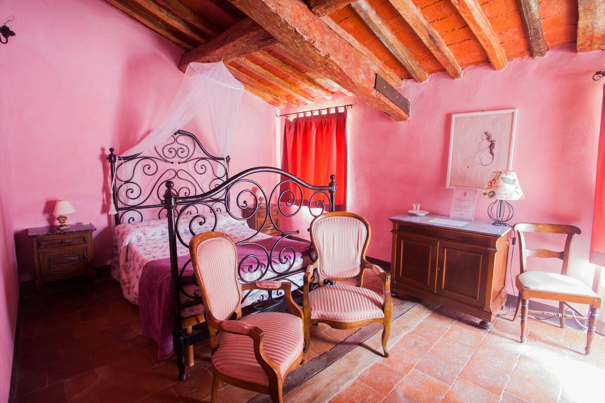 Ein Schlafzimmer mit einem Himmelbett, zwei Holzstühlen, einem Schreibtisch aus Holz und einem Nachttisch mit Lampe.